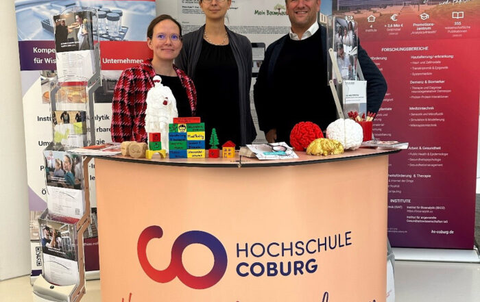 Drei Vertreter präsentieren hinter einem Promotion-Tisch Lehrmaterialien und Spielsachen der Hochschule Coburg. Im Hintergrund geben zwei große Banner Einblicke in die Welt der Bioanalytik.