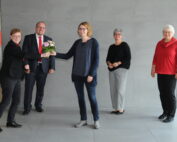 Fünf Menschen stehen gemeinsam vor einer grauen Wand in einem Innenbereich der Hochschule Coburg. Eine Person im schwarzen Outfit überreicht einer anderen Person im blauen Pullover einen Blumenstrauß. Drei weitere, zwei Frauen und ein Mann, lächeln und beobachten die Szene. Im Hintergrund sind Stühle zu sehen.