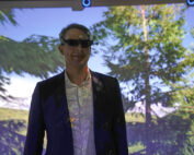 Ein Mann in dunklem Anzug und weißem Hemd steht vor einem Bildschirm, auf dem eine malerische Naturansicht mit Bäumen und Himmel an der Hochschule Coburg zu sehen ist. Er trägt eine spezielle 3D-Brille und die Szene wird auf ihn projiziert, wodurch eine bunte Überlagerung entsteht.