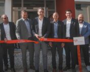 Eine Gruppe von acht Männern in Anzügen steht vor dem Gebäude der Hochschule Coburg und hält bei einer feierlichen Eröffnung ein rotes Band in der Hand. Der Mann in der Mitte schneidet das Band an diesem sonnigen Tag im Freien mit einer großen Schere durch.