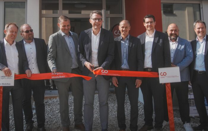Eine Gruppe von acht Männern in Anzügen steht vor dem Gebäude der Hochschule Coburg und hält bei einer feierlichen Eröffnung ein rotes Band in der Hand. Der Mann in der Mitte schneidet das Band an diesem sonnigen Tag im Freien mit einer großen Schere durch.