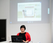 Eine Person in einem roten Pullover steht an einem Rednerpult mit einem Computer und hält einen Vortrag vor einem Publikum an der Hochschule Coburg. Der Raum hat weiße Wände und eine geschlossene schwarze Tür, während die projizierte Folie auf dem Bildschirm ein Bild eines Tablets und verschiedener Gegenstände auf einem Tisch zeigt.