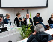 Ein fünfköpfiges Gremium der Hochschule Coburg sitzt vor einem Publikum in einem Hörsaal. Eine Frau gestikuliert beim Sprechen, während die anderen aufmerksam zuhören. Auf einem Tisch in der Nähe stehen Wasserflaschen, daneben ein geöffneter Laptop. Im Vordergrund steht eine Pflanze.
