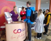 Am Informationsstand der Hochschule Coburg herrscht reges Treiben, während zwei Vertreter mit den Besuchern sprechen. Dahinter informieren Broschüren und eine Schautafel vor einem leuchtend roten und orangefarbenen Hintergrund über die Studienfächer.