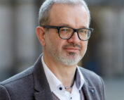 Ein Mann mit kurzen grauen Haaren und Brille, der eine graue Jacke, ein weißes Hemd und eine Weste trägt, steht im Freien vor einem unscharfen Hintergrund. Sein gelassenes Auftreten spiegelt seine Verbindung zur Hochschule Coburg wider.