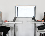 Zwei Menschen mit Masken sitzen einander gegenüber an getrennten Schreibtischen mit Laptops. Zwischen ihnen ist auf einem Bildschirm ein Flussdiagramm zu sehen. Die Szenerie ist ein moderner, heller Büroraum der Hochschule Coburg.