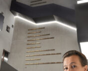 Eine Frau steht in einem modernen, betonierten Innenraum der Hochschule Coburg, umgeben von eckigen Treppen und Kunstausstellungen. Sie blickt in die Kamera, eingerahmt von einem gut beleuchteten Raum und künstlerischen Installationen mit linearen Elementen.