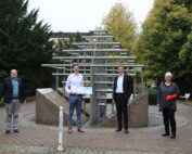 Vier Personen stehen draußen neben einer Metallskulptur in einer Parkanlage der Hochschule Coburg. Drei Männer in Business-Kleidung und eine Frau mit einem roten Gipsverband am Arm posieren für ein Gruppenfoto. Im Hintergrund sind Bäume und die Ecke eines Gebäudes zu sehen, die die lebendige Campus-Atmosphäre einfangen.