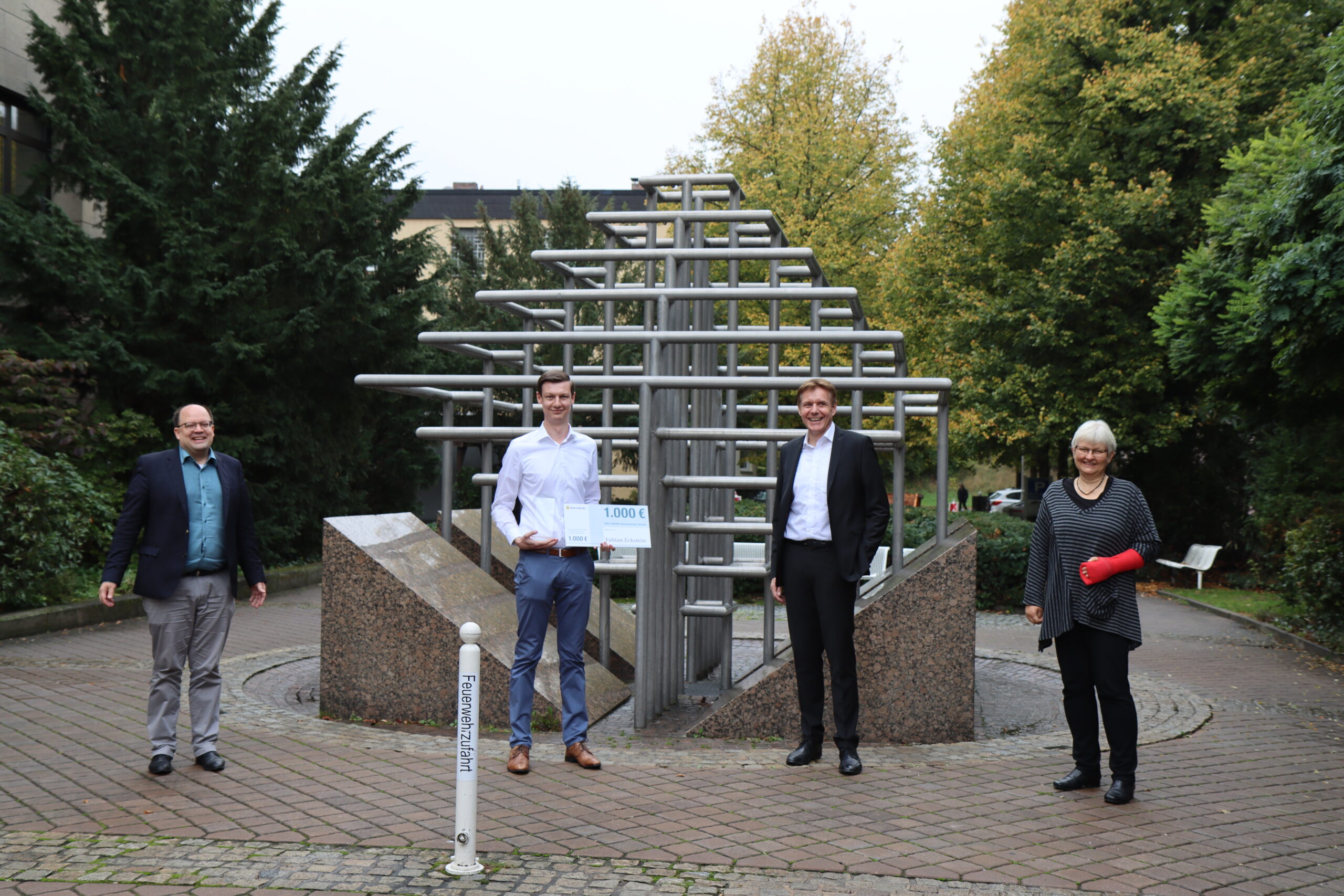 Vier Personen stehen draußen neben einer Metallskulptur in einer Parkanlage der Hochschule Coburg. Drei Männer in Business-Kleidung und eine Frau mit einem roten Gipsverband am Arm posieren für ein Gruppenfoto. Im Hintergrund sind Bäume und die Ecke eines Gebäudes zu sehen, die die lebendige Campus-Atmosphäre einfangen.