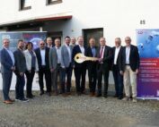 Eine Gruppe von fünfzehn Personen in formeller Kleidung steht vor einem Gebäude und lächelt für ein Foto. Zwei Personen in der Mitte halten einen großen symbolischen Schlüssel, der die Hochschule Coburg repräsentiert. Auf beiden Seiten der Gruppe sind Banner mit Text zu sehen.