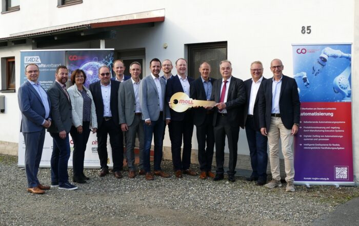 Eine Gruppe von fünfzehn Personen in formeller Kleidung steht vor einem Gebäude und lächelt für ein Foto. Zwei Personen in der Mitte halten einen großen symbolischen Schlüssel, der die Hochschule Coburg repräsentiert. Auf beiden Seiten der Gruppe sind Banner mit Text zu sehen.