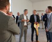 Eine Gruppe von Männern in Anzügen steht in einem geräumigen, unfertigen Raum der Hochschule Coburg. Ein Mann hält einen großen dekorativen Schlüssel in der Hand und sie scheinen in ein Gespräch vertieft zu sein, möglicherweise bei einer Präsentation oder Veranstaltung.