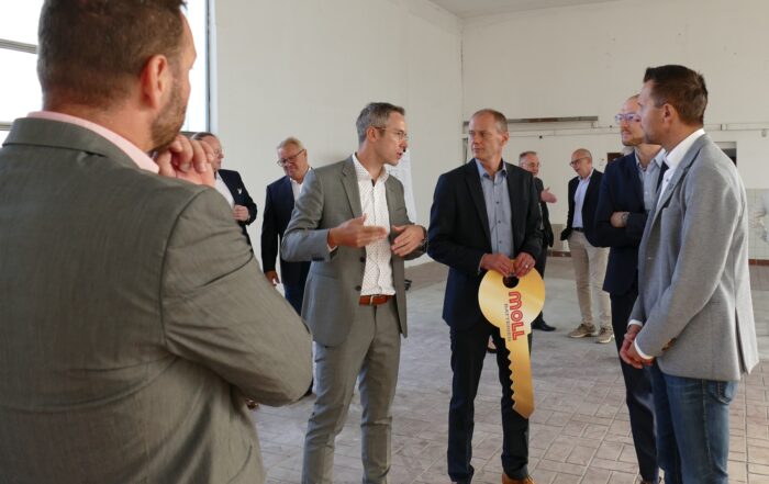 Eine Gruppe von Männern in Anzügen steht in einem geräumigen, unfertigen Raum der Hochschule Coburg. Ein Mann hält einen großen dekorativen Schlüssel in der Hand und sie scheinen in ein Gespräch vertieft zu sein, möglicherweise bei einer Präsentation oder Veranstaltung.
