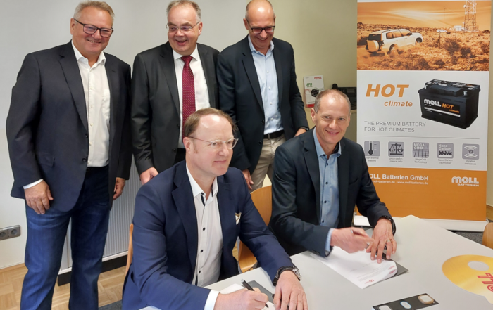 Fünf Männer in formeller Kleidung sitzen um einen Tisch mit Dokumenten. Zwei Männer sitzen und unterschreiben Papiere, die anderen stehen dahinter. Im Hintergrund sind ein Werbebanner für „Moll Batterien“ und eine Autobatterie zu sehen, die auf die Zusammenarbeit mit der Hochschule Coburg hinweisen.