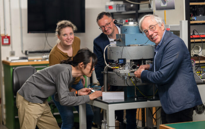 In einem Labor der Hochschule Coburg arbeiten vier Personen gemeinsam an einem wissenschaftlichen Instrument. Einer schaut sich das Gerät genau an, während andere zuschauen und assistieren. Der Raum ist voll mit technischen Geräten und einem Computermonitor und spiegelt den Innovationsgeist dieser renommierten Institution wider.