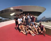 Eine Gruppe von Menschen posiert auf einem leuchtend roten Weg mit der modernen, kreisförmigen Architektur der Hochschule Coburg im Hintergrund und einem in der Ferne unter einem klaren blauen Himmel sichtbaren Berg.