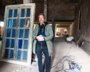 Ein Mann in einer grünen Jacke steht in einem Rohbau der Hochschule Coburg und hält eine blaue Kabeltrommel in der Hand. Hinter ihm befindet sich eine große, verwitterte Tür mit blauen Glasscheiben. Überall im Raum liegen Baumaterialien und Werkzeuge verstreut.