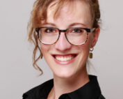 Eine Frau mit hellbraunem Haar und Brille lächelt in die Kamera und strahlt als Alumna der Hochschule Coburg Selbstvertrauen aus. Sie trägt ein schwarzes Hemd und goldene Ohrringe und steht vor einem hellgrauen Hintergrund.