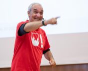Ein Mann mit kurzen grauen Haaren und einem roten T-Shirt mit weißem Logo spricht in ein Headset-Mikrofon. Er strahlt, zeigt nach vorne und steht selbstbewusst vor einer weißen Leinwand in der Hochschule Coburg.