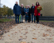 Eine Gruppe von sechs Personen steht auf einem mit Herbstlaub geschmückten Kiesweg, einer hält einen Blumenstrauß in der Hand. Sie befinden sich im Freien in der Nähe der Hochschule Coburg, hinter ihnen ein großes Gebäude und an den Seiten Bäume.