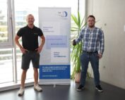Zwei Männer stehen drinnen neben einem Roll-up-Banner und verkörpern damit den Innovationsgeist und die Zusammenarbeit mit der Hochschule Coburg. Der Mann links trägt ein schwarzes Poloshirt und Shorts, während sein Gegenüber sich für ein kariertes Hemd und Jeans entscheidet.