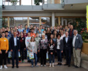 Eine große Gruppe von Menschen, darunter Erwachsene und Jugendliche, hat sich für ein Foto in einem Innenraum der Hochschule Coburg versammelt. Einige halten Geschenktüten in den Händen. Im Hintergrund ist ein modernes Interieur mit Pflanzen und Glaswänden zu sehen.