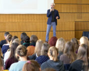 Ein Redner steht auf der Bühne der Hochschule Coburg und spricht zu einem aufmerksamen Publikum im Hörsaal. Der Raum ist mit Holzpaneelen und einer großen Projektionsleinwand ausgestattet, was die spannende Atmosphäre verstärkt, während sich die unterschiedlichsten Teilnehmer auf die Präsentation konzentrieren.