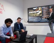 In einem modernen Konferenzraum der Hochschule Coburg diskutieren drei Männer über eine Präsentation auf einem großen Bildschirm. Ein Mann steht und zeigt auf den Bildschirm, die anderen beiden sitzen auf einer roten Couch, vor ihnen steht ein aufgeklappter Laptop.