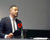 Ein Mann in Anzug und Krawatte steht hinter einem Tisch und hält ein Tablet mit dem Logo der Hochschule Coburg in der Hand. Auf dem Tisch liegen eine rote Blume in einer kleinen Glasflasche und einige Papiere. Im Hintergrund ist eine leere Wand.