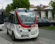 Ein autonomer Shuttlebus in den rot-weißen Farben der Hochschule Coburg fährt auf einer Kopfsteinpflasterstraße. In der Nähe parkt ein schwarzer Bus, eingerahmt von den grünen Bäumen und städtischen Gebäuden im Hintergrund.