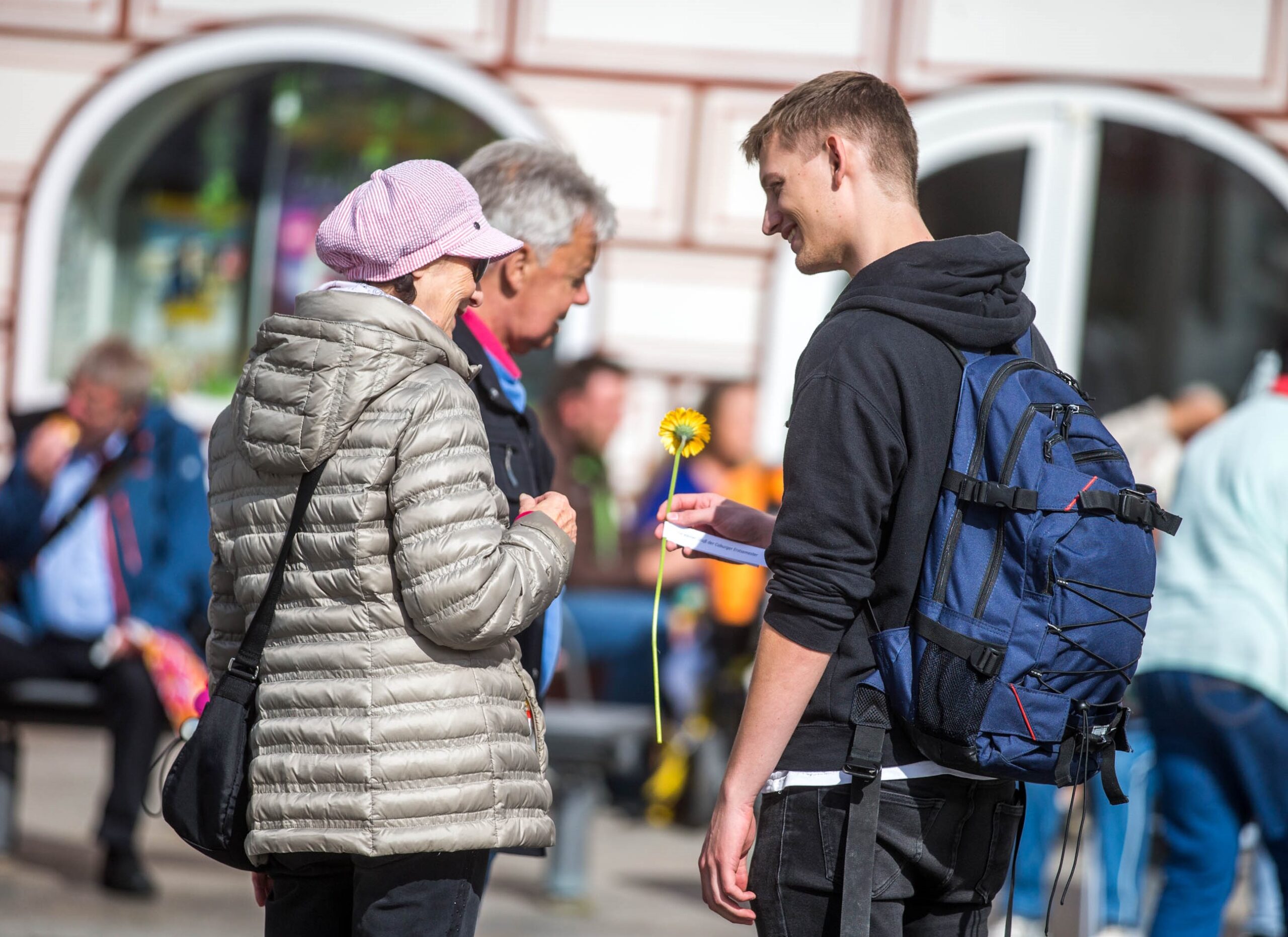 Ein junger Mann mit einem Rucksack der Hochschule Coburg lächelt, als er einer älteren Frau in leichter Jacke und rosafarbenem Hut eine gelbe Blume überreicht. In der Nähe beobachtet ein Mann diese freundliche Geste im lebhaften öffentlichen Raum im Hintergrund, in dem sich viele Menschen tummeln.