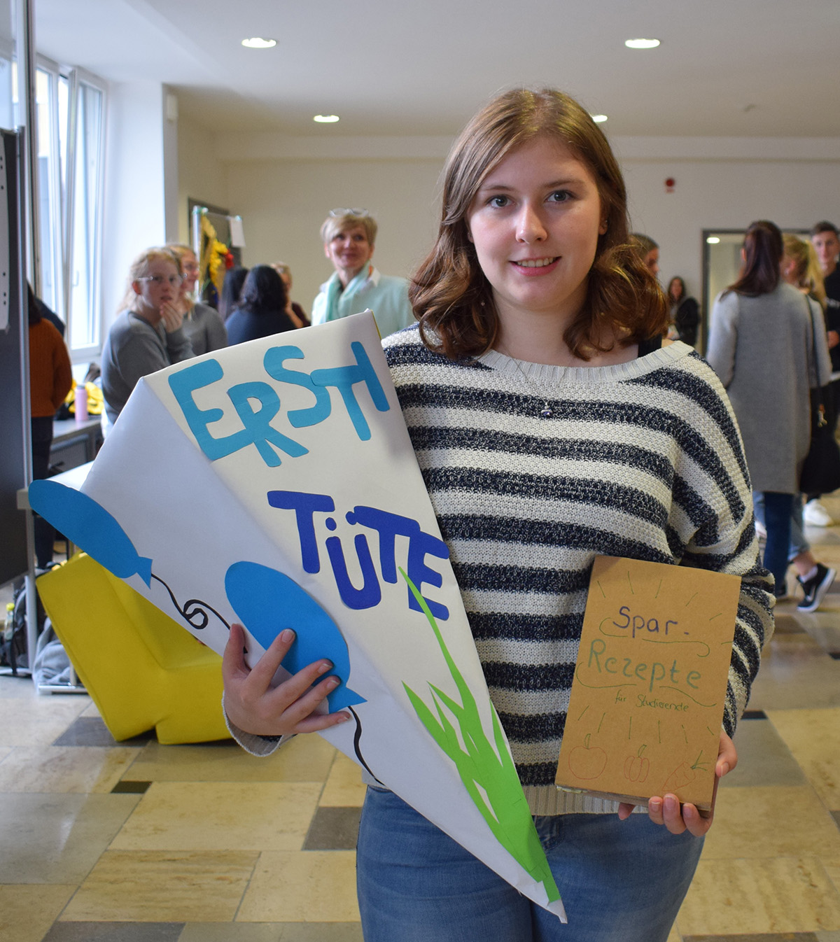 Eine Frau steht drinnen und hält eine große Papiertüte mit der Aufschrift „Erst Tüte“. Außerdem hält sie eine braune Papiertüte mit der Aufschrift „Spar Rezepte“, vielleicht Teil eines Kreativprojekts der Hochschule Coburg. Im Hintergrund sind Menschen zu sehen.