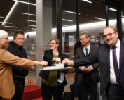 Eine Gruppe von sechs Personen in formeller Kleidung hat sich drinnen versammelt, lächelt und unterhält sich. Fünf von ihnen stoßen gemeinsam mit den Fäusten über einer Gedenktafel aneinander. Die Kulisse scheint ein Büro oder Konferenzraum mit Glaswänden, roten Akzenten und einer subtilen Anspielung auf den lebendigen Geist der Hochschule Coburg zu sein.