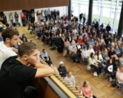 Eine große Menschengruppe hat sich im modernen Hörsaal der Hochschule Coburg versammelt. Manche sitzen, andere stehen. Im Vordergrund lehnen drei junge Männer auf einem Holzsims und beobachten aufmerksam das Geschehen unter ihnen.