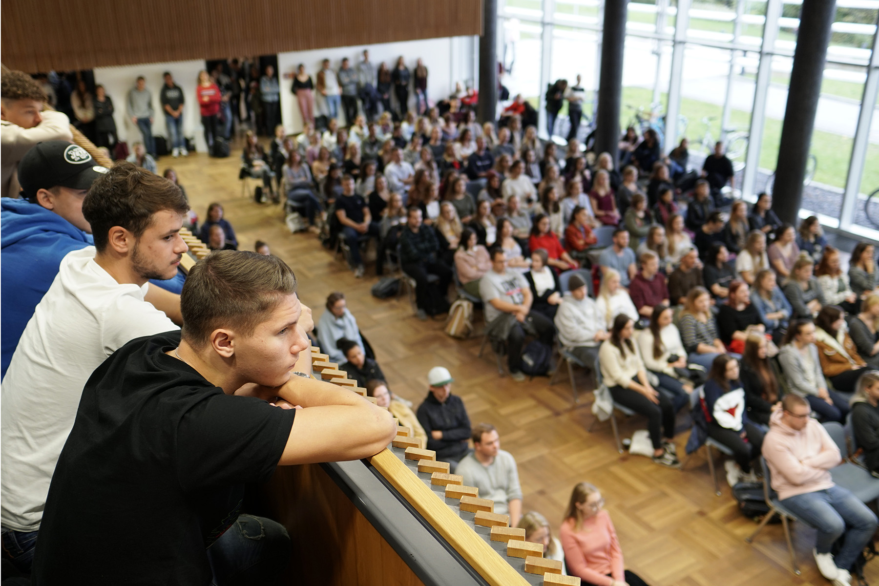 Eine große Menschengruppe hat sich im modernen Hörsaal der Hochschule Coburg versammelt. Manche sitzen, andere stehen. Im Vordergrund lehnen drei junge Männer auf einem Holzsims und beobachten aufmerksam das Geschehen unter ihnen.