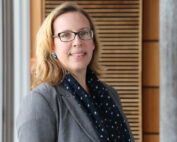 Eine Person mit schulterlangem Haar und Brille trägt einen grauen Blazer und einen dunklen Schal mit Sternenmuster. Sie steht im Innenbereich neben den Holzlatten eines Gebäudes der Hochschule Coburg und lächelt herzlich in die Kamera.