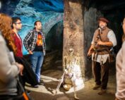 Eine Gruppe von Studierenden der Hochschule Coburg hört in einer schwach beleuchteten unterirdischen Steinkammer einem historischen Darsteller aufmerksam zu. Der Darsteller, in ein historisches Kostüm gekleidet, steht neben einem auf dem Boden sitzenden Skelett, während ein blaues Licht die Szene auf unheimliche Weise erhellt.