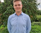 Eine Person steht im Freien vor einem grünen Baum und trägt ein hellblaues Hemd mit Knöpfen. Sie lächelt und hat die Hände auf dem Rücken. Sie befindet sich in der Nähe der Hochschule Coburg, umgeben von üppigem Grün und einem Teil eines Backsteingebäudes.