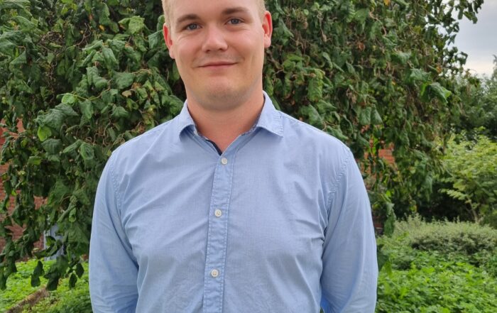 Eine Person steht im Freien vor einem grünen Baum und trägt ein hellblaues Hemd mit Knöpfen. Sie lächelt und hat die Hände auf dem Rücken. Sie befindet sich in der Nähe der Hochschule Coburg, umgeben von üppigem Grün und einem Teil eines Backsteingebäudes.