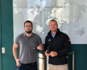 Zwei Männer stehen drinnen vor einer Weltkartenausstellung. Ein Mann, der eine Brille und ein graues Poloshirt trägt, hält einen kleinen Gegenstand in der Hand. Der andere, der eine schwarze Jacke der Hochschule Coburg trägt, hält ebenfalls einen ähnlichen Gegenstand in der Hand. Sie lächeln in die Kamera.