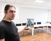 Eine Person mit langen Haaren, einem schwarzen Hemd und einem Hochschulabzeichen der Hochschule Coburg steht in einem Konferenzraum und hält ein Tablet mit dem Bild einer Statue in der Hand. Im Raum stehen ein langer weißer Tisch und schwarze Stühle, im Hintergrund ist teilweise ein Banner zu sehen.