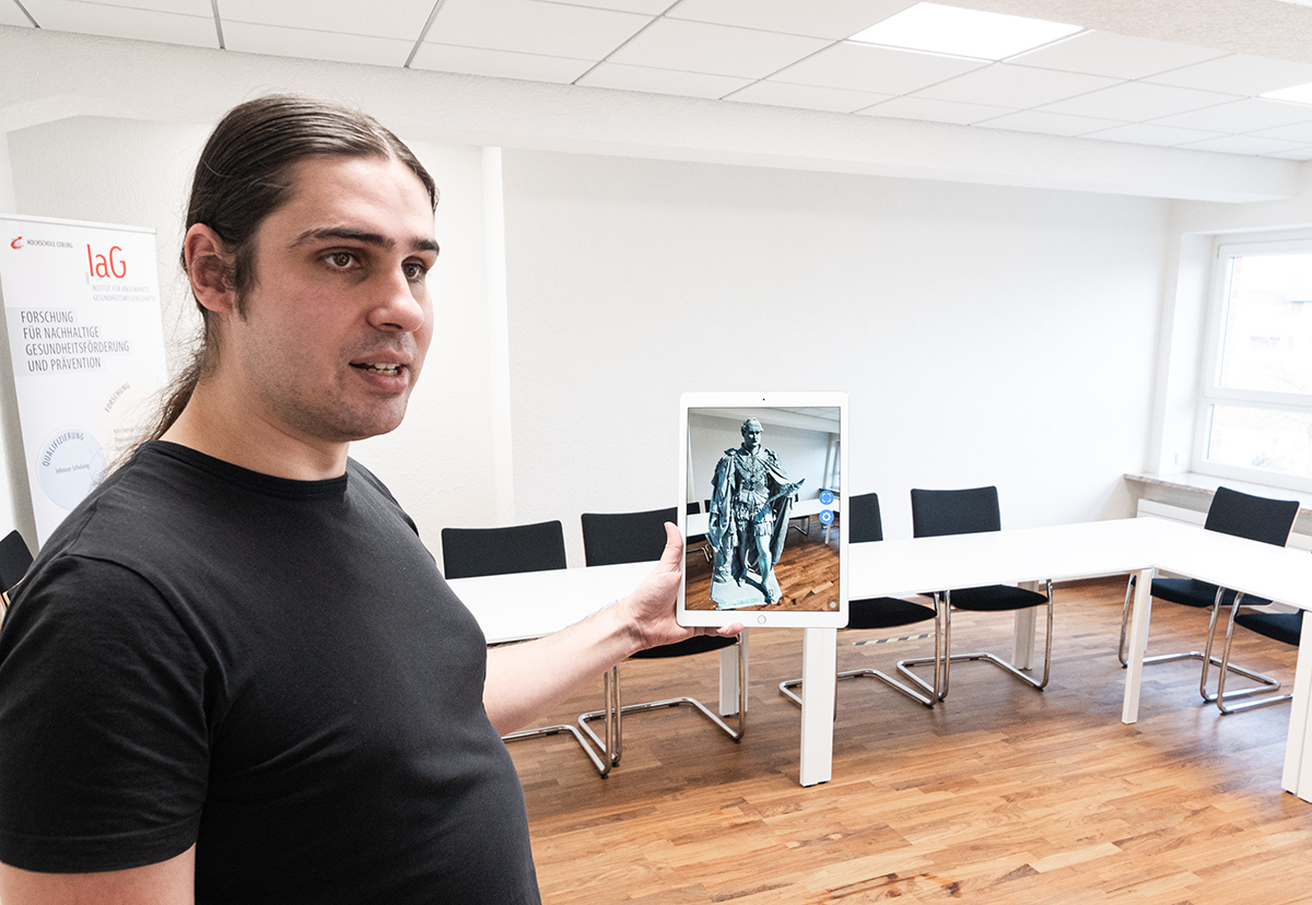 Eine Person mit langen Haaren, einem schwarzen Hemd und einem Hochschulabzeichen der Hochschule Coburg steht in einem Konferenzraum und hält ein Tablet mit dem Bild einer Statue in der Hand. Im Raum stehen ein langer weißer Tisch und schwarze Stühle, im Hintergrund ist teilweise ein Banner zu sehen.