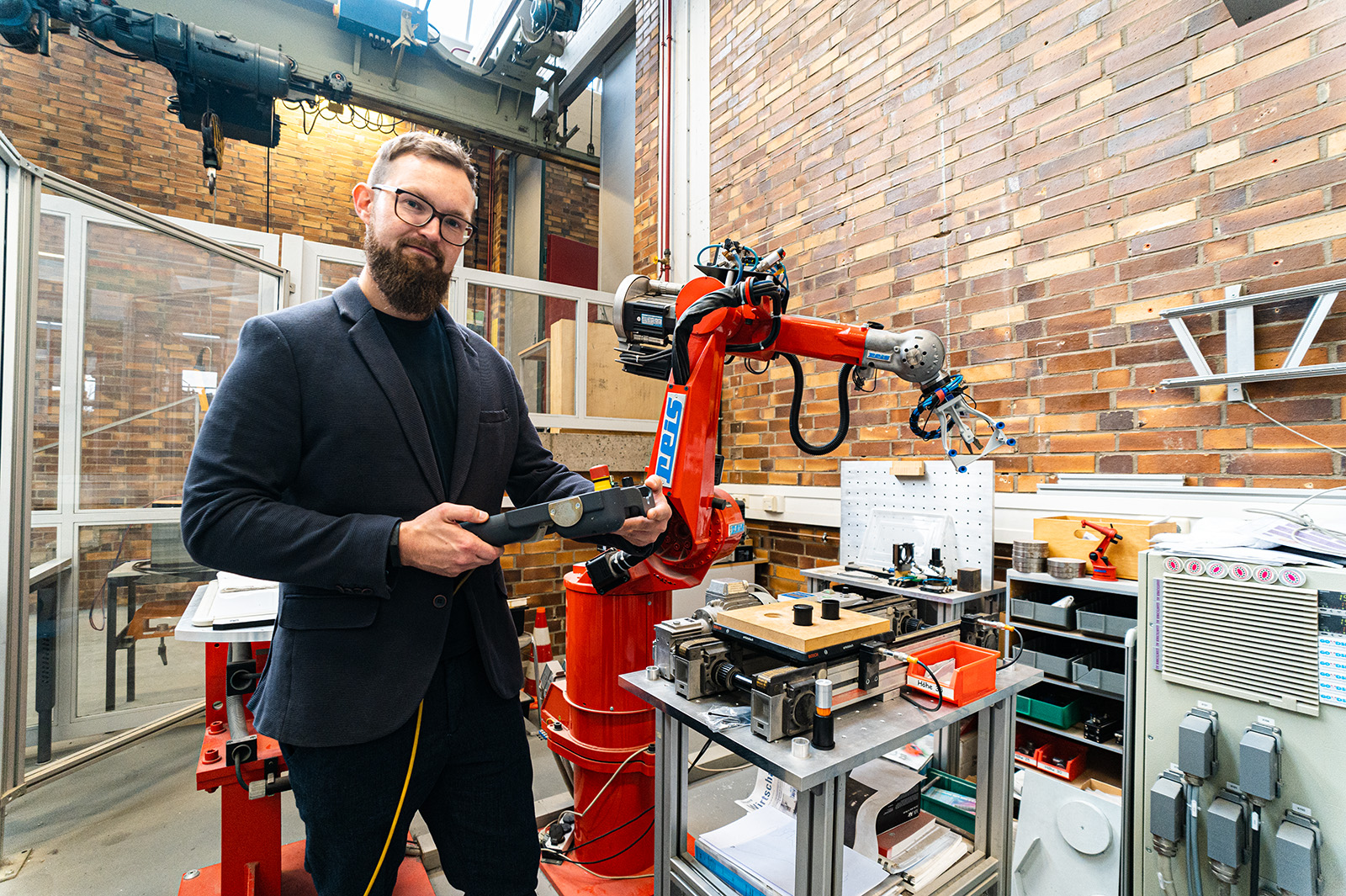 Eine Person mit Bart, Brille und schwarzer Jacke steht neben einem großen roten Roboterarm in einer Industrieanlage der Hochschule Coburg. Sie hält ein Werkzeug und lächelt. Im Hintergrund sind Backsteinmauern und verschiedene Geräte und Regale zu sehen.