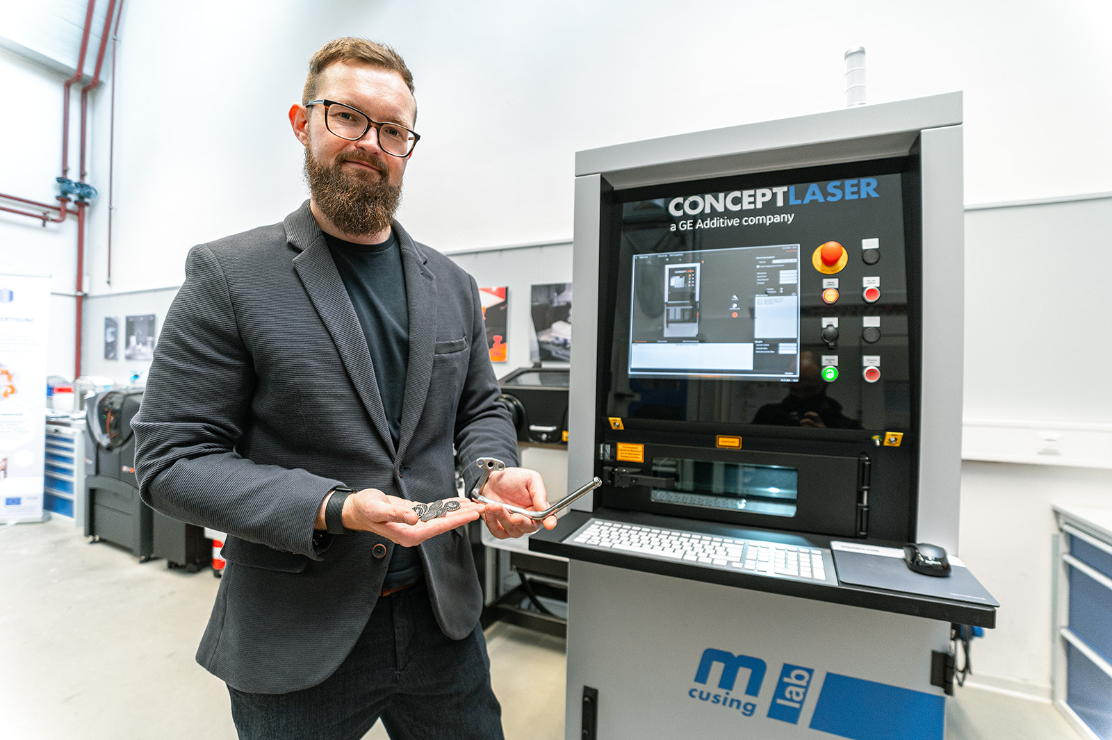 Ein Mann mit Bart und Brille steht vor einem 3D-Drucker der Hochschule Coburg und hält zwei Metallteile in der Hand. Er trägt einen dunklen Blazer und Jeans, auf dem Display des 3D-Druckers sind verschiedene Bedienelemente und Einstellungen zu sehen.