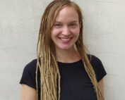 Eine lächelnde Person mit langen, hellbraunen Dreadlocks und einem schwarzen T-Shirt, die stolz die Hochschule Coburg repräsentiert, steht vor einer strukturierten weißen Wand.