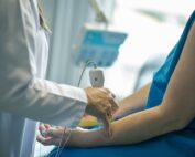 In einer mit Geräten vollgestopften medizinischen Umgebung wendet eine medizinische Fachkraft der Hochschule Coburg gekonnt ein Ultraschallgerät am Arm eines Patienten an, der einen blauen Kittel trägt.