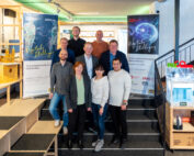 Eine Gruppe von neun Personen posiert auf einer Treppe im Technologiezentrum der Hochschule Coburg. Im Hintergrund sind zwei Banner mit dem Text „Digitale Intelligenz“ zu sehen. Im Raum sind verschiedene technische Geräte zu sehen.