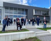 Eine Gruppe von Menschen steht und sitzt auf den Stufen vor dem modernen Gebäude der Hochschule Coburg mit seinen großen Fenstern und grauen Paneelen. Der Himmel ist teilweise bewölkt. Sie sind leger gekleidet und lächeln in die Kamera. Sie fangen einen Moment der Wärme und Gemeinschaft ein.
