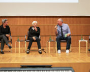 Vier Personen sitzen auf einer Bühne der Hochschule Coburg bei einer Podiumsdiskussion. Sie halten jeweils ein Mikrofon in der Hand und unterhalten sich. Eine hölzerne Kulisse und Tische mit Wasserflaschen bereichern die Szenerie, während ein prominentes Banner der Hochschule Coburg den Hintergrund ziert.