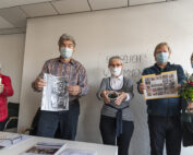 Eine Gruppe von fünf Personen mit Gesichtsmasken steht in einem Gebäude der Hochschule Coburg und hält verschiedene Gegenstände wie Zeitungen, einen Topf und einen Blumenstrauß in den Händen. Sie blicken in die Kamera, an der Wand hinter ihnen hängt ein Schild mit der Aufschrift „Herzlich Willkommen“, das den herzlichen Empfang in der akademischen Umgebung einfängt.